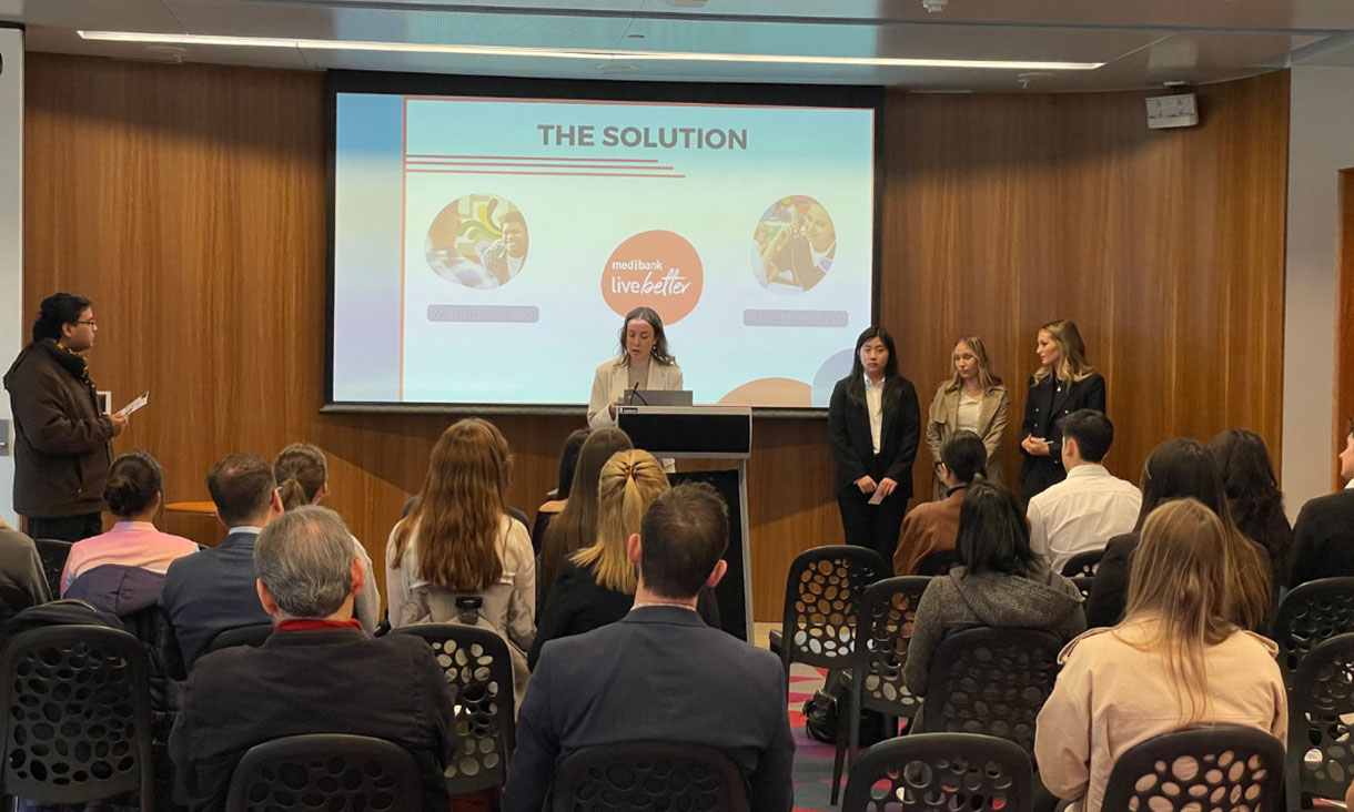 Students sit on chairs watching a project team present at a lectern in front of a projection screen.