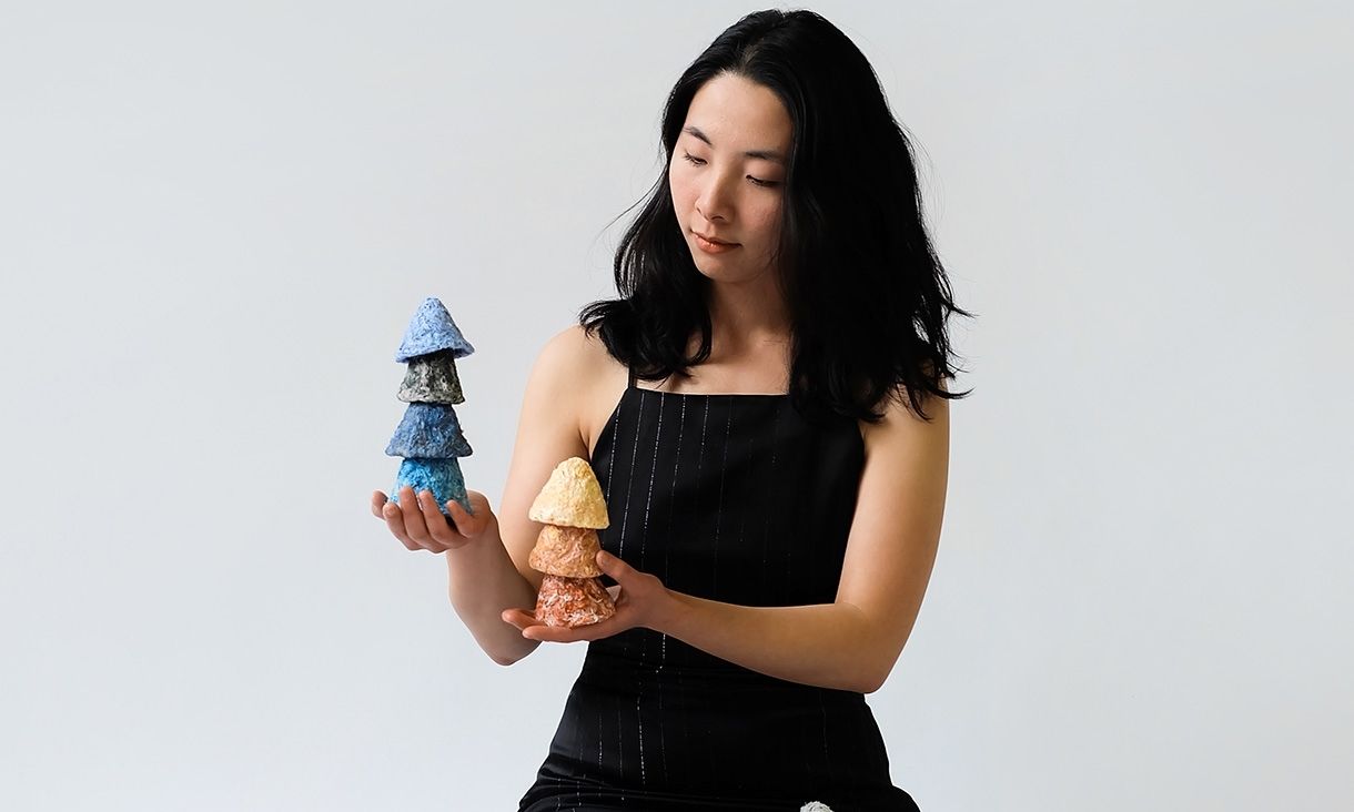 Photography of artist Vivian Qiu in black dress holding two small sculptures in her hands. 