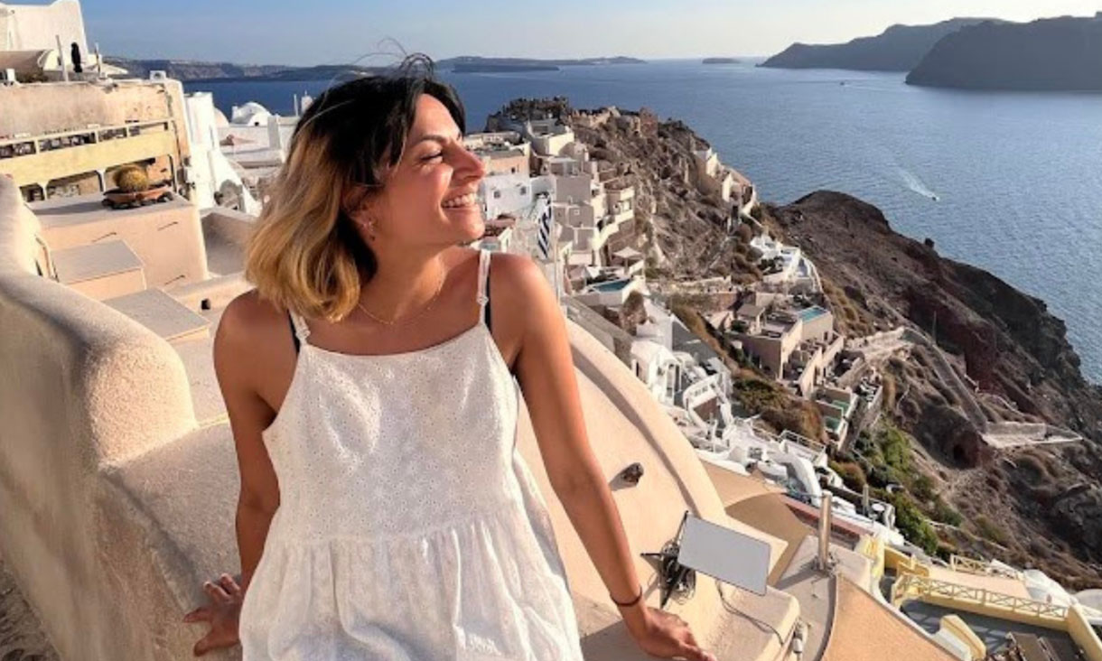 Woman in white dress standing in front of a cliff with water in the background.