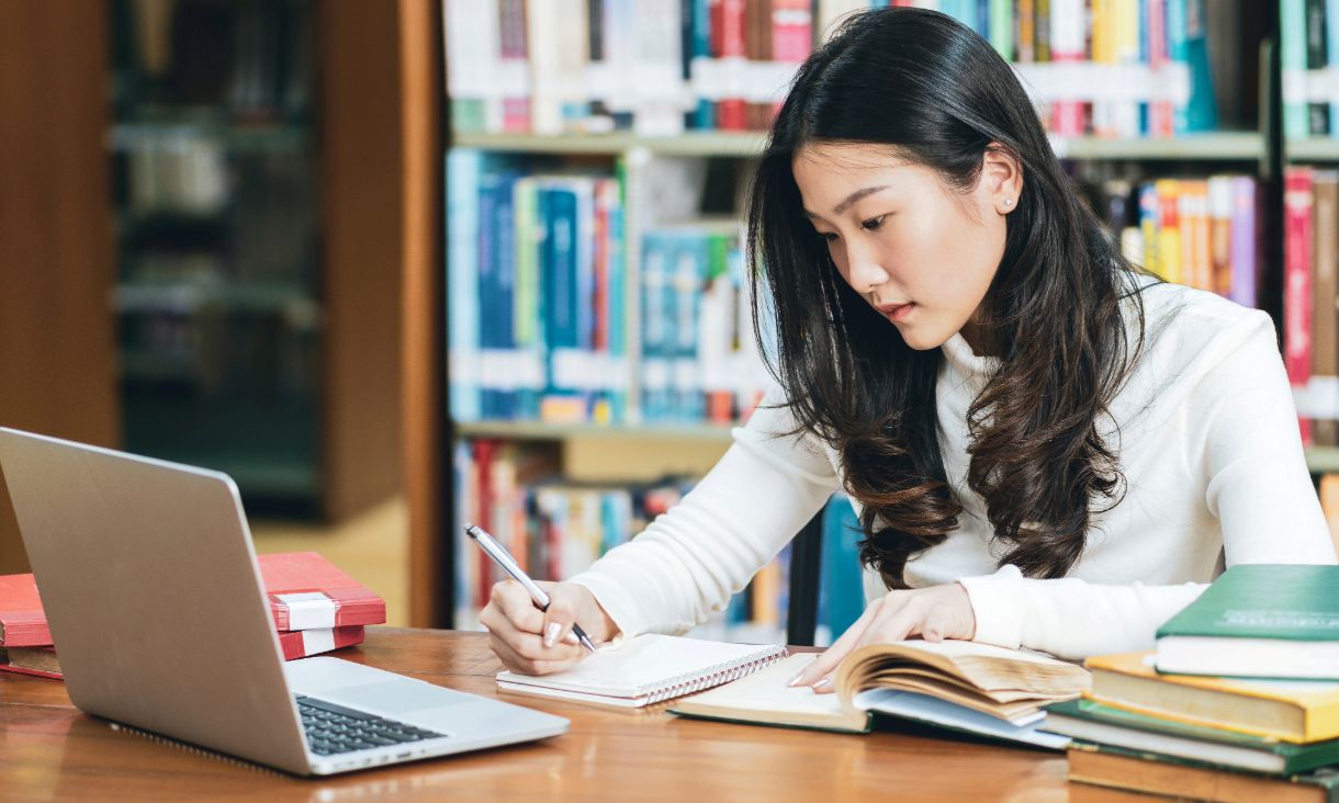 Stock image: Woman at a desk writing on a notepad Surrounded by books and a laptop.