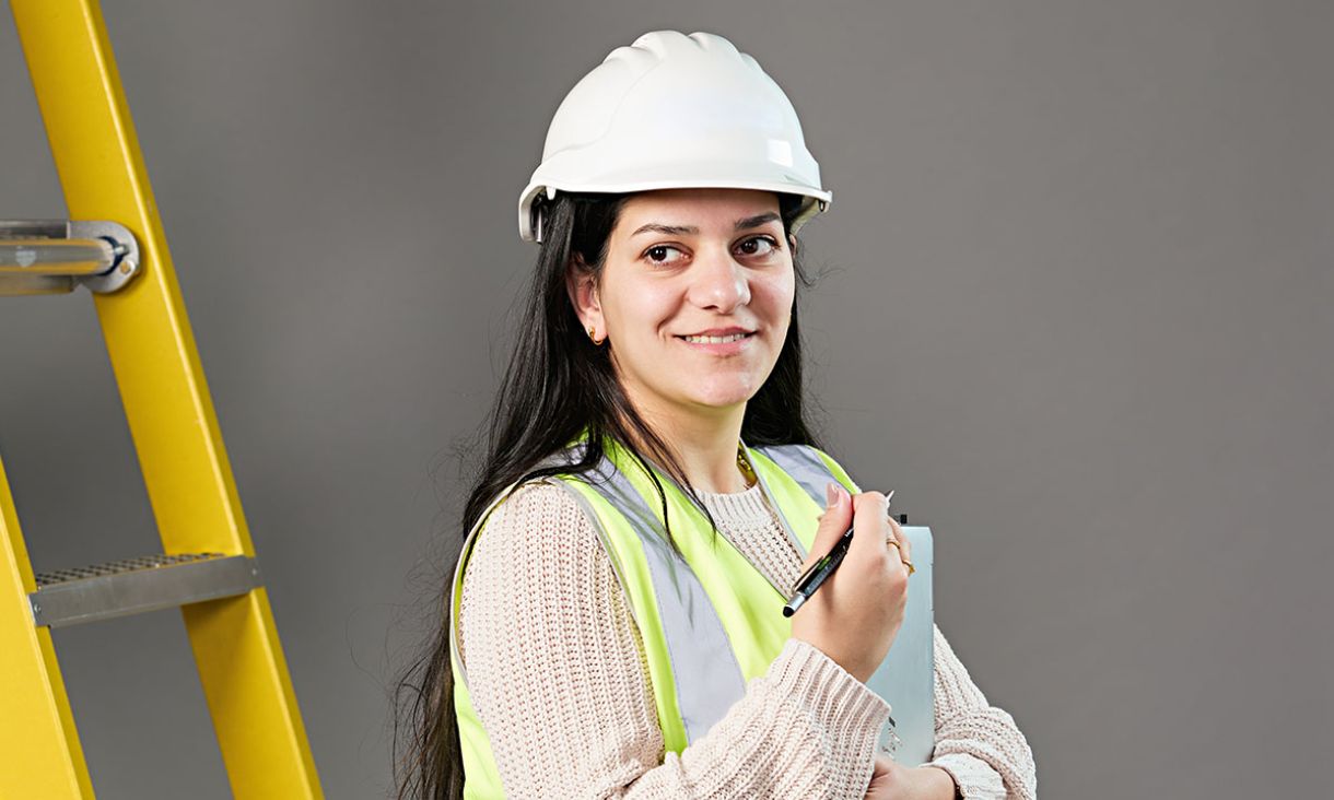 A woman wearing a hard hat and safety vest, holding a clipboard, stands in a construction environment.