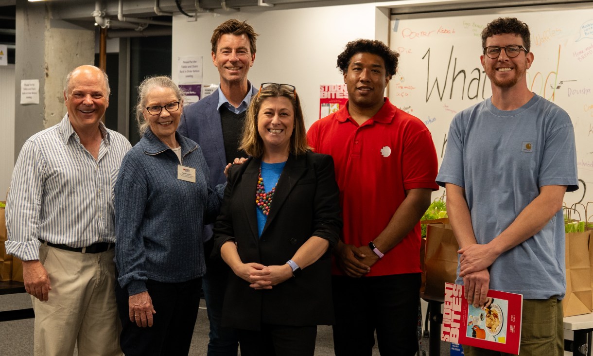 Six people stand together in a room with tables of packed grocery bags behind them at the RMIT Eats free groceries initiative.