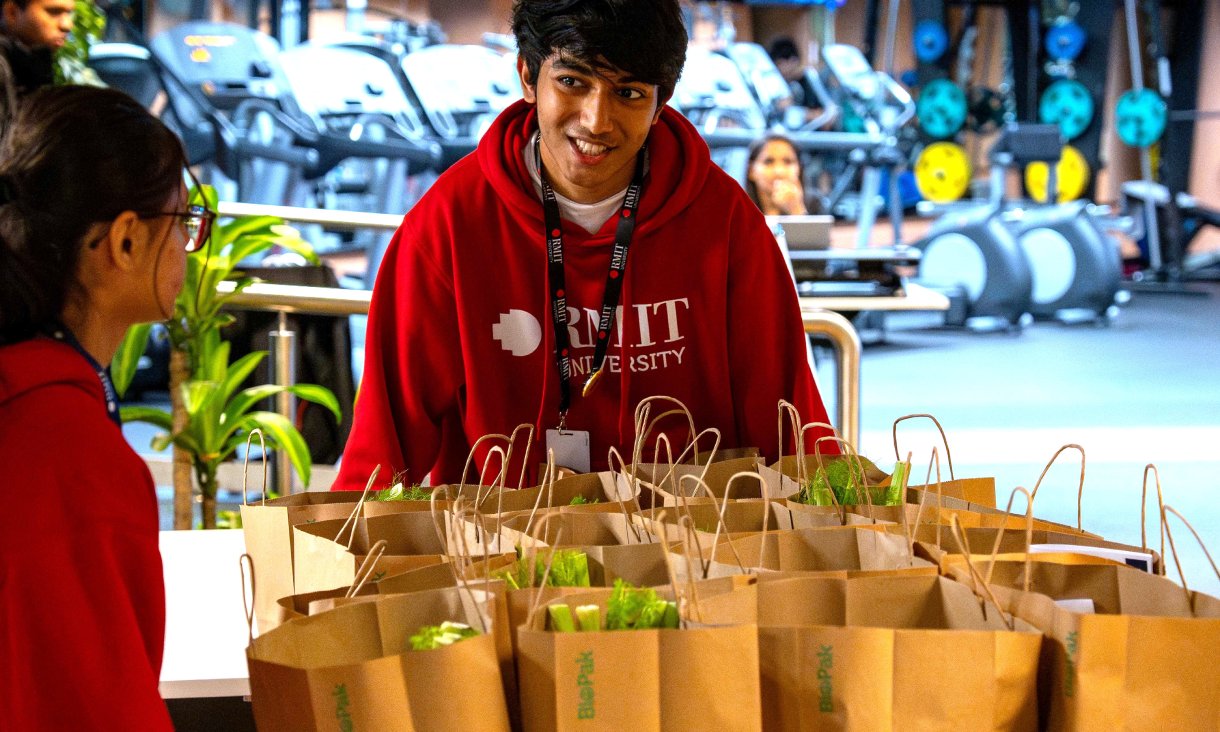 Two people in red RMIT hoodies stand beside a table filled with packed grocery bags at the RMIT Eats initiative, with gym equipment in the background.