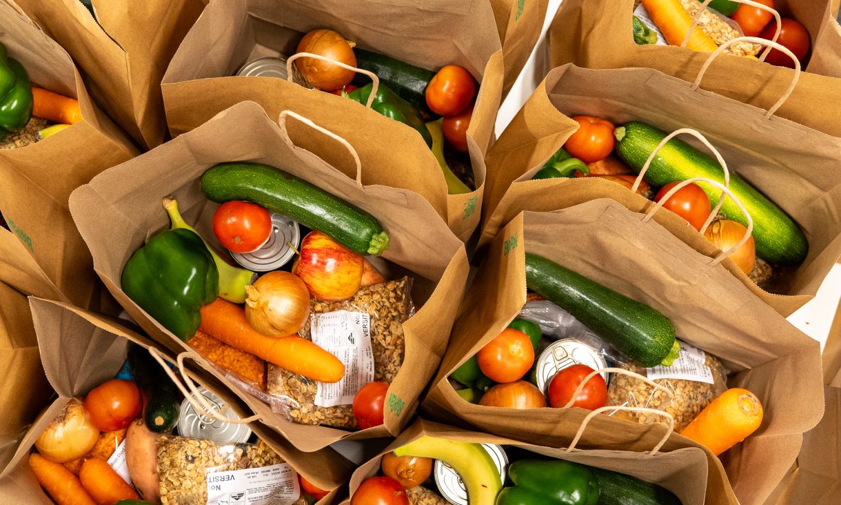 Paper grocery bags filled with fresh produce and pantry items, including zucchini, capsicum, carrots, tomatoes, bananas and canned food.