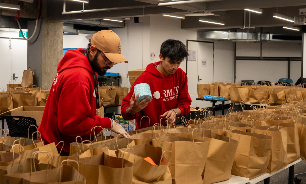 Two people in red RMIT hoodies pack grocery bags in a large room lined with tables covered in brown paper bags.