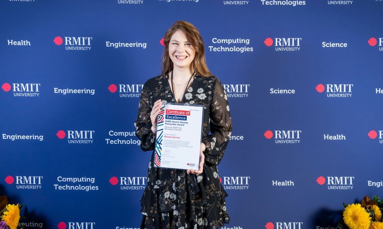 Student standing in front of RMIT media wall smiling while holding an award