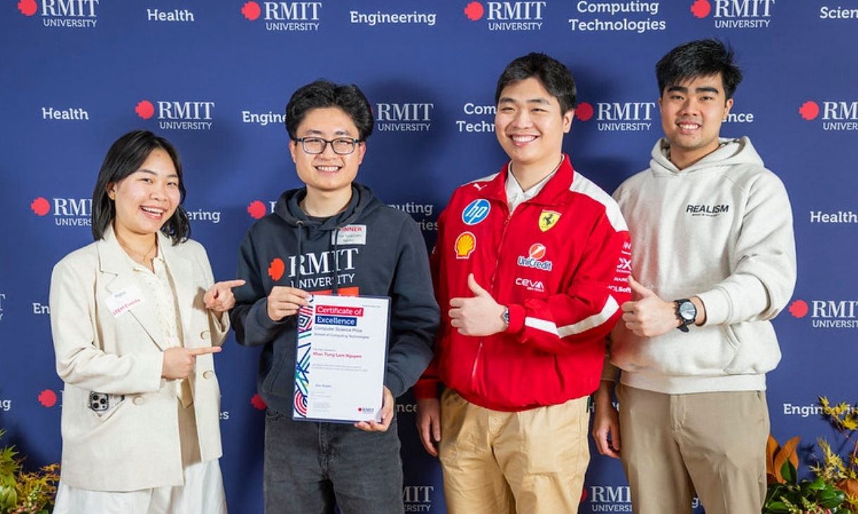Four students standing in front of media wall smiling while holding award