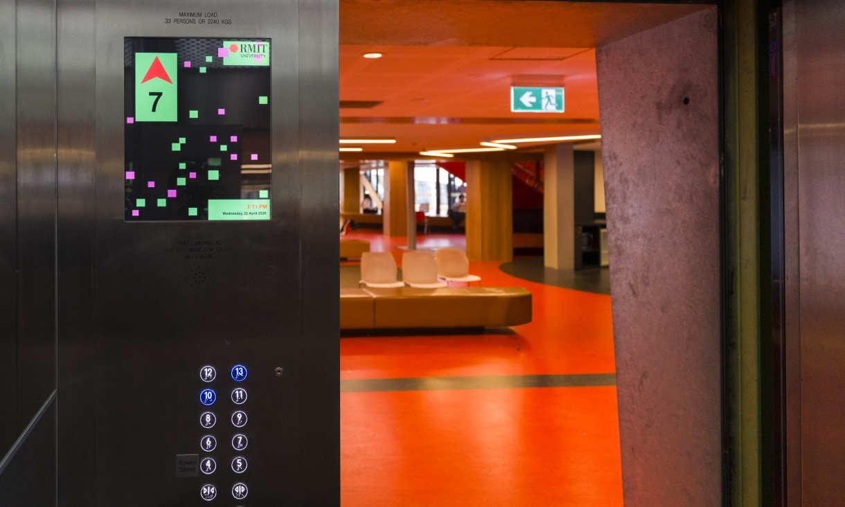 Interior of a lift in Building 10 at RMIT, showing a digital screen with graphic elements from Artificial Intelligelism and the building interior visible through the open lift doorway.