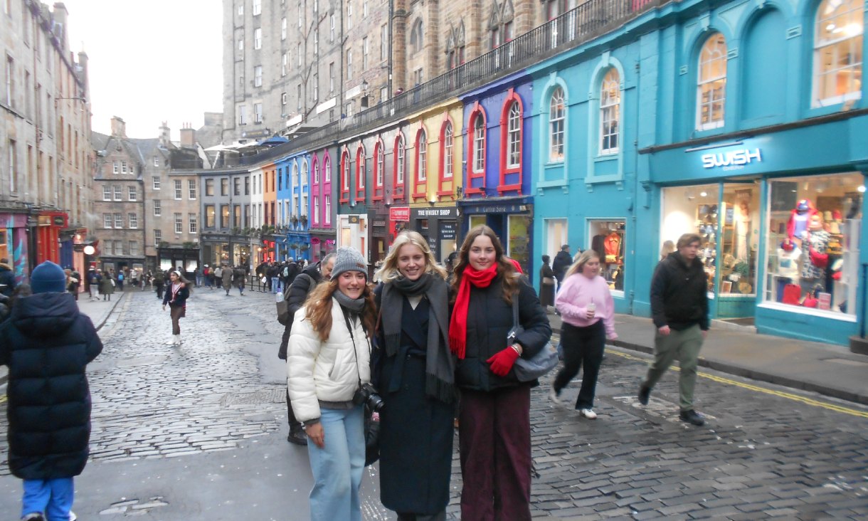 Three people standing on the cobblestones of Victoria Street in Edinburgh, with colourful shopfronts and historic stone buildings behind them.