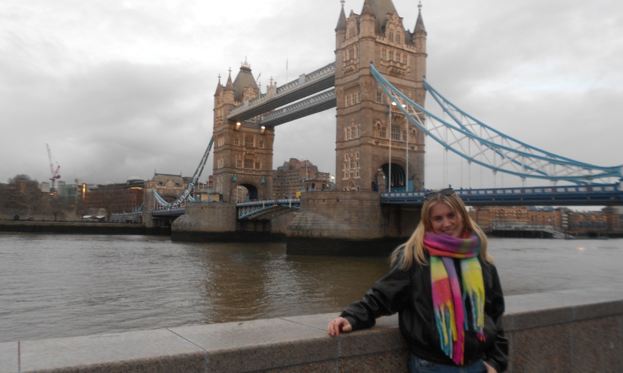 One person standing beside the River Thames with Tower Bridge in the background under an overcast sky.