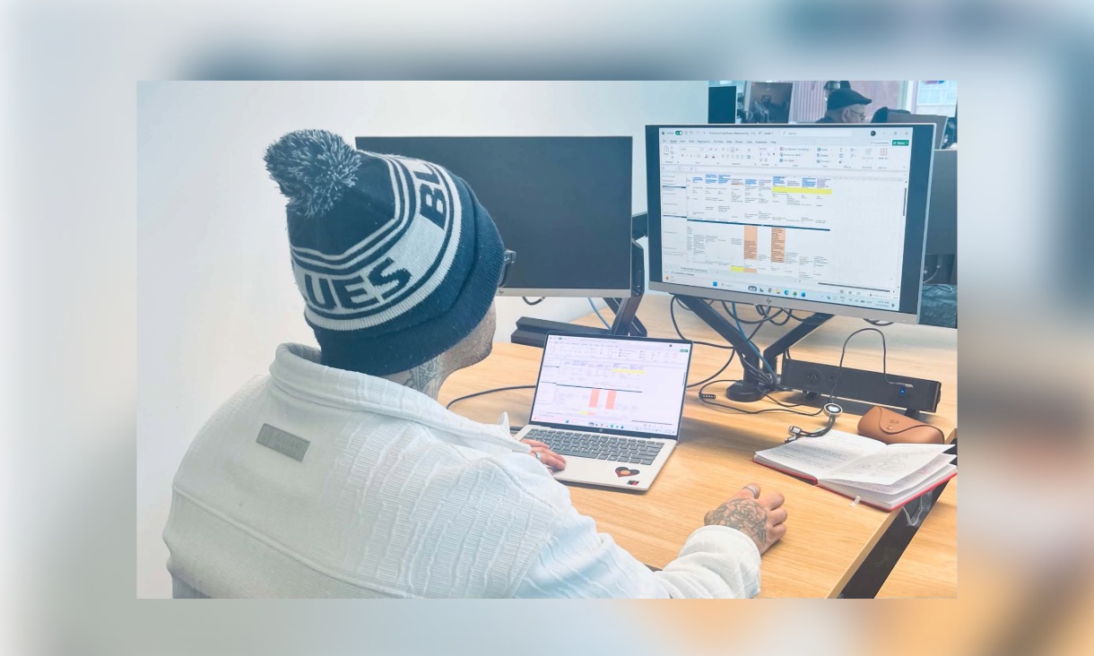 Dillon Stocks in a beanie working on a spreadsheet at a desk with a laptop and monitor.