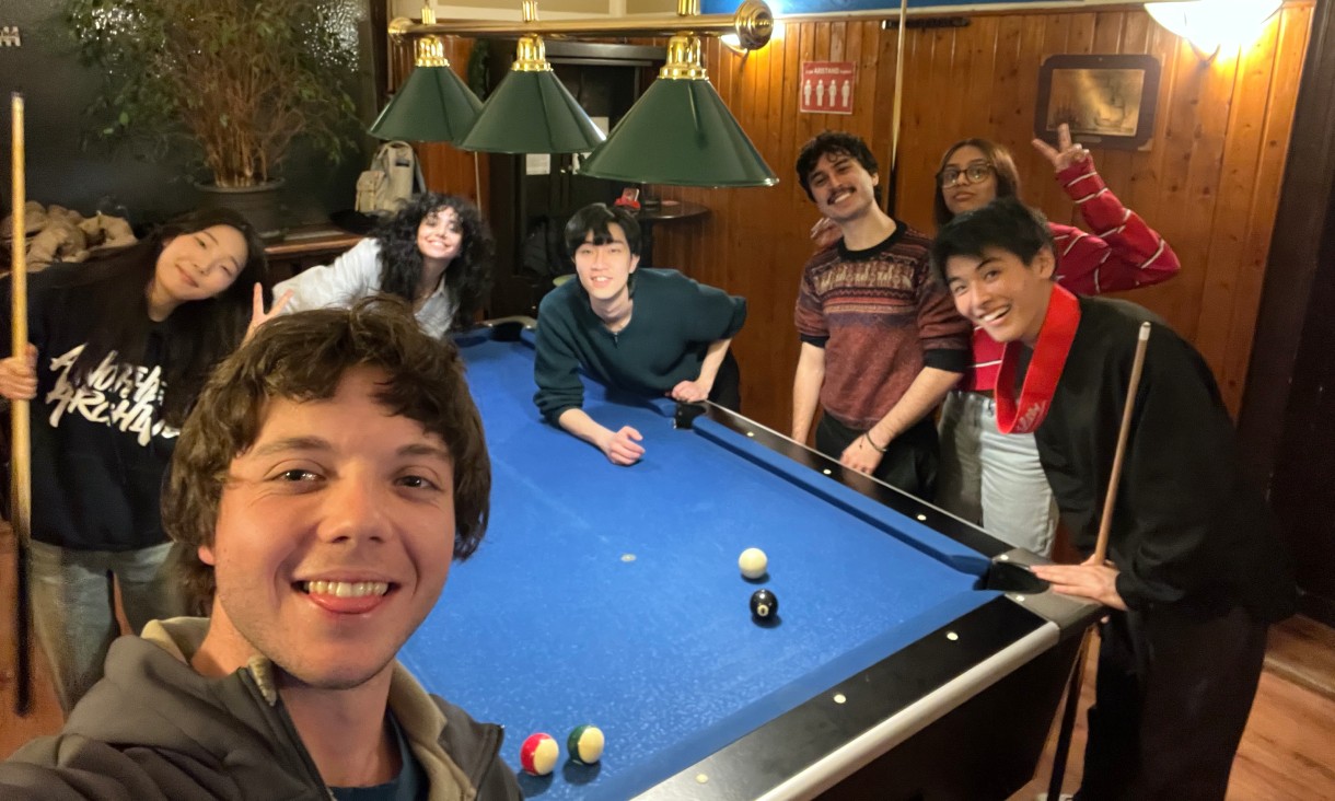 Seven people gathered around a blue pool table in a wood-panelled indoor venue, with pool cues and balls visible.