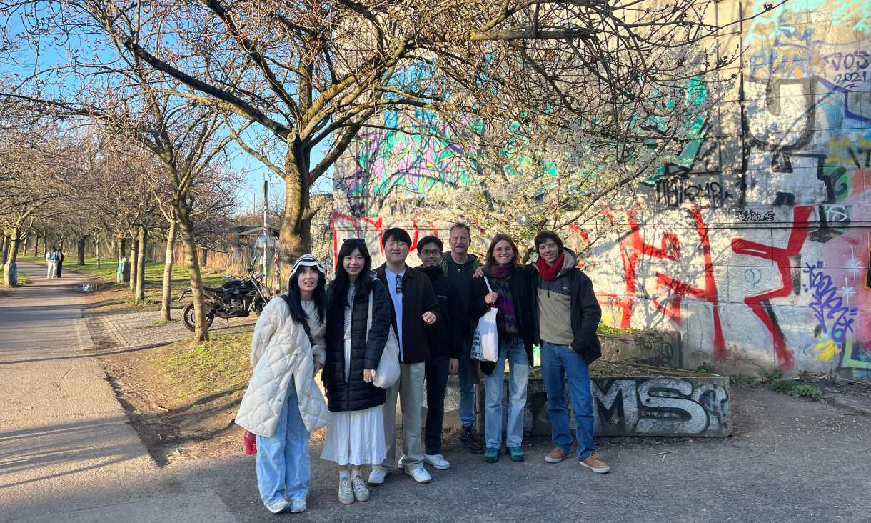 Seven people standing together beside a graffiti-covered wall and a tree-lined path on a sunny day.