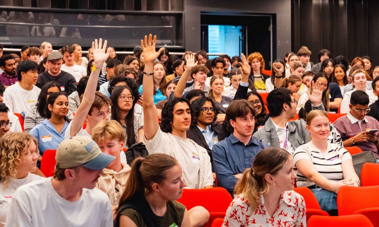 Students seated in a packed lecture theatre raise their hands during a presentation at the Student Leadership Summit.