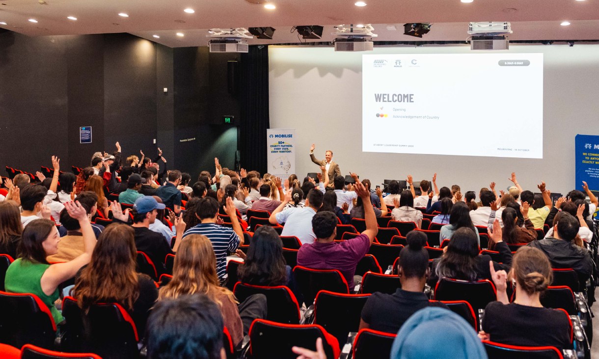 Students seated in a packed lecture theatre raise their hands while a presenter speaks at the front of the room during the Student Leadership Summit.