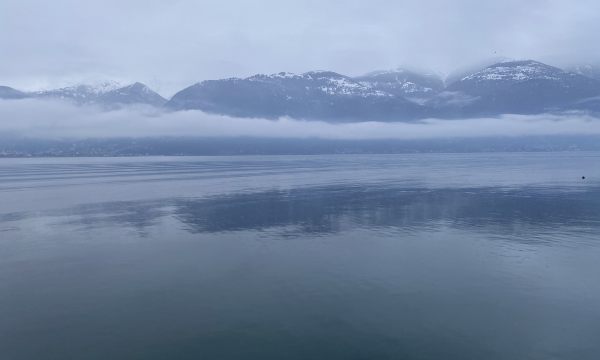 Calm lake water reflecting a cloudy sky, with low clouds and snow-capped mountains along the opposite shore.