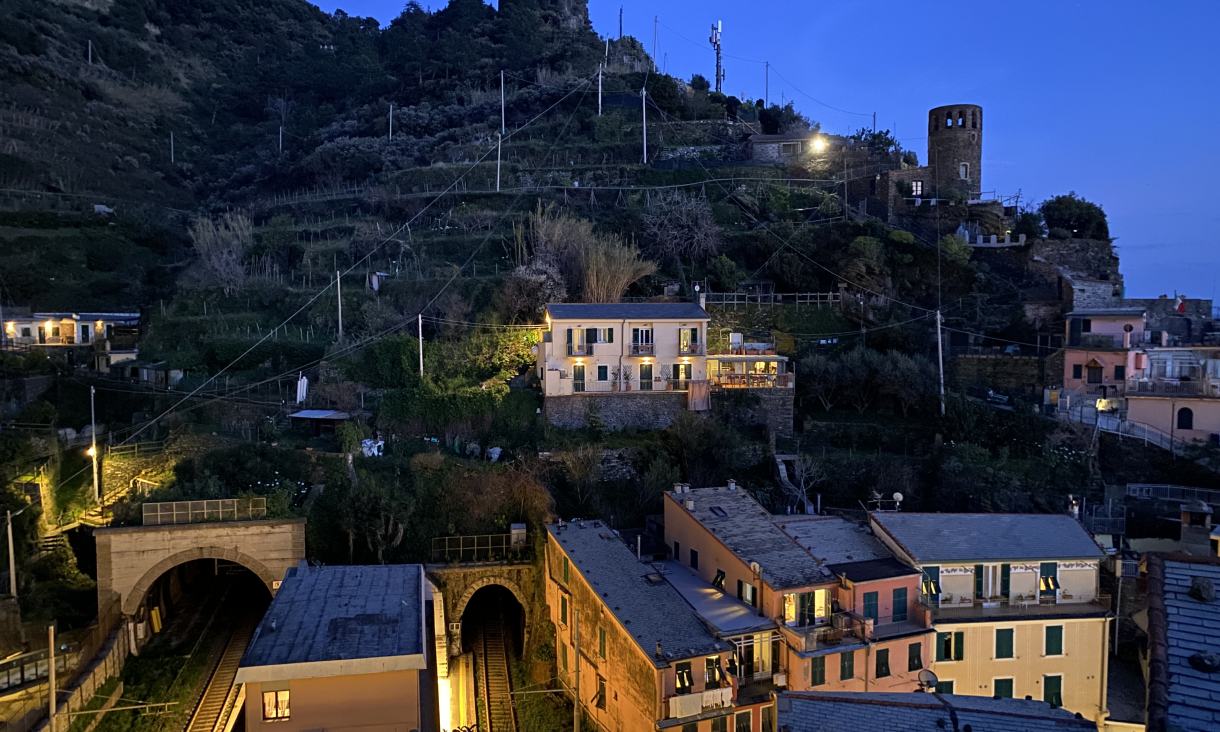 Railway tunnels and colourful buildings beneath a terraced hillside and stone tower at dusk in Monterosso al Mare.