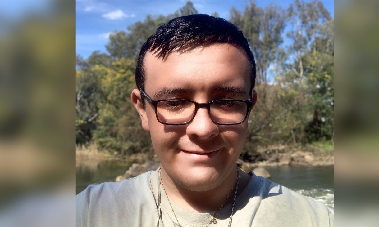 Tobi Ward smiling in a selfie by a river on a sunny day, with gum trees behind him.