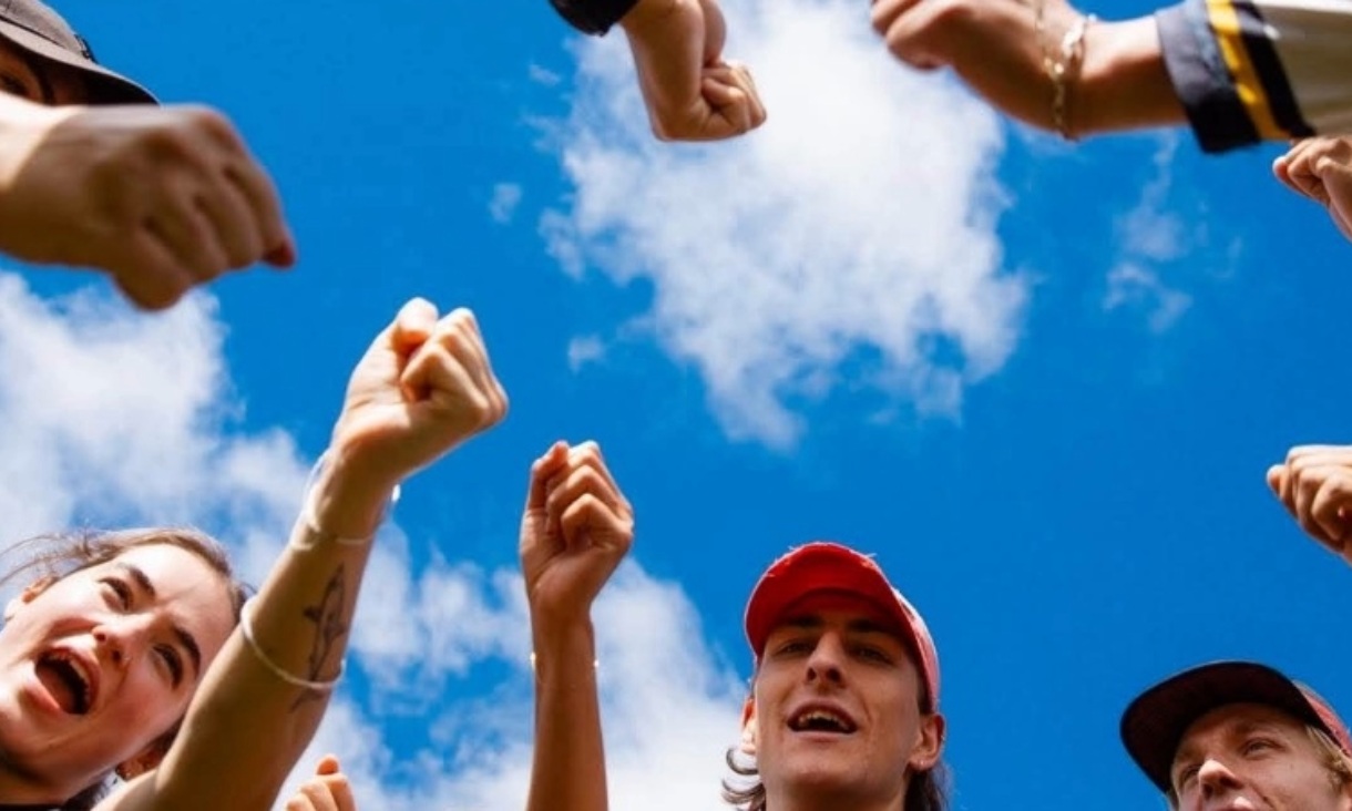 Looking up at a circle of young people raising their fists towards a bright blue sky, some smiling and cheering.