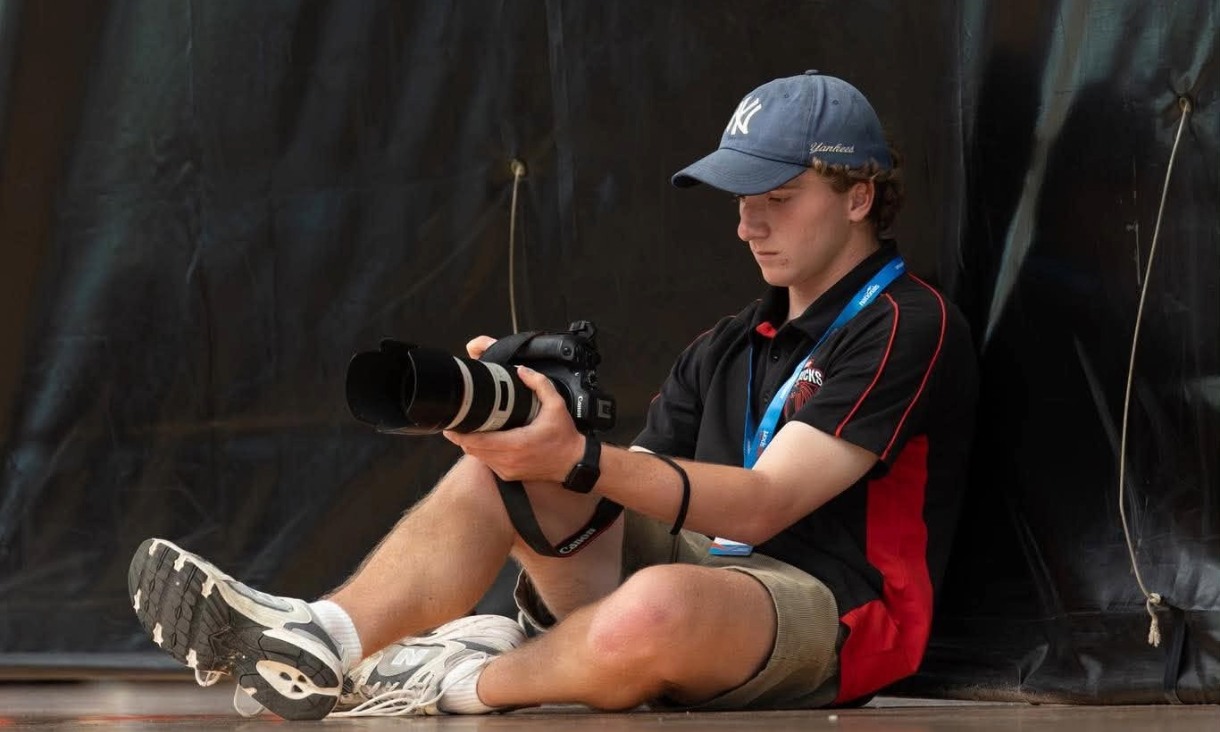A young man in a black and red polo shirt and cap sits against a dark curtain, looking at the screen of his camera.