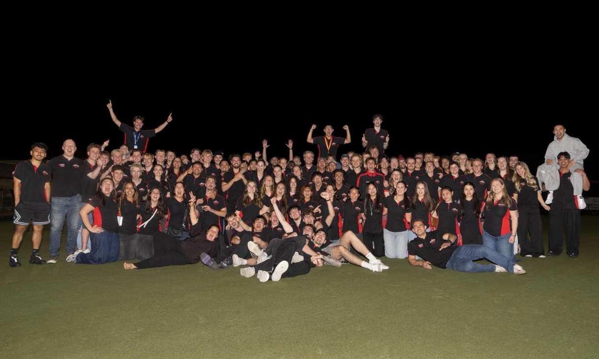 A large group in matching black and red polo shirts gathers on a grass field at night, cheering with arms raised.