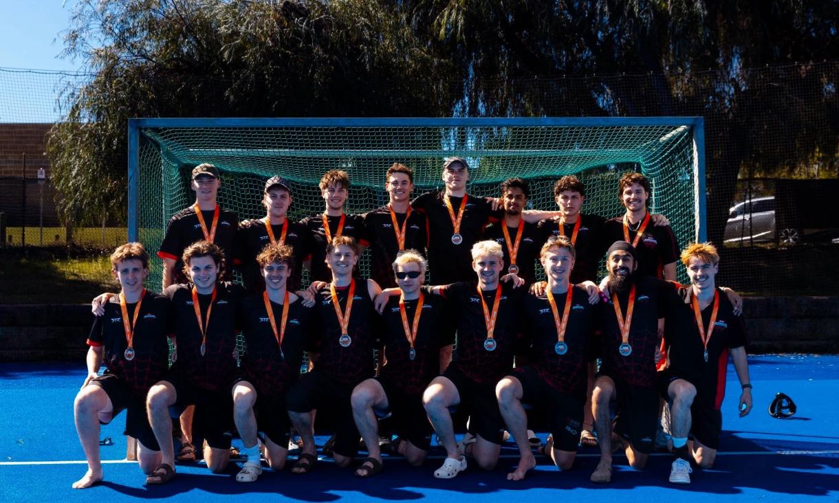 A hockey team in black and red uniforms poses in two rows in front of a goal on a blue pitch, each wearing a silver medal.