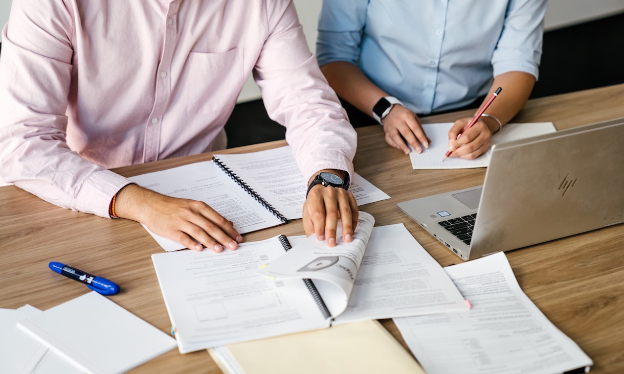 Two students work on paperwork and laptops.