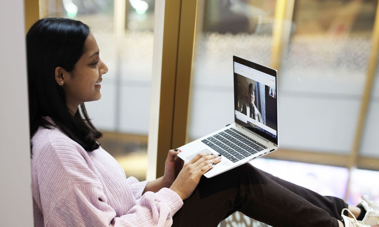 A girl with dark hair in a pink jumper smiles at someone on a laptop screen.