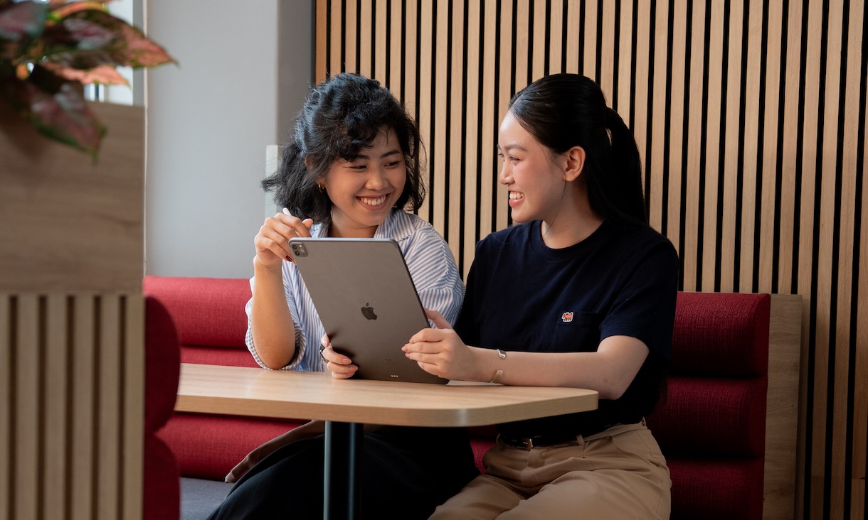 Two students sit together in a modern study space, smiling as they look at a tablet device on the table.