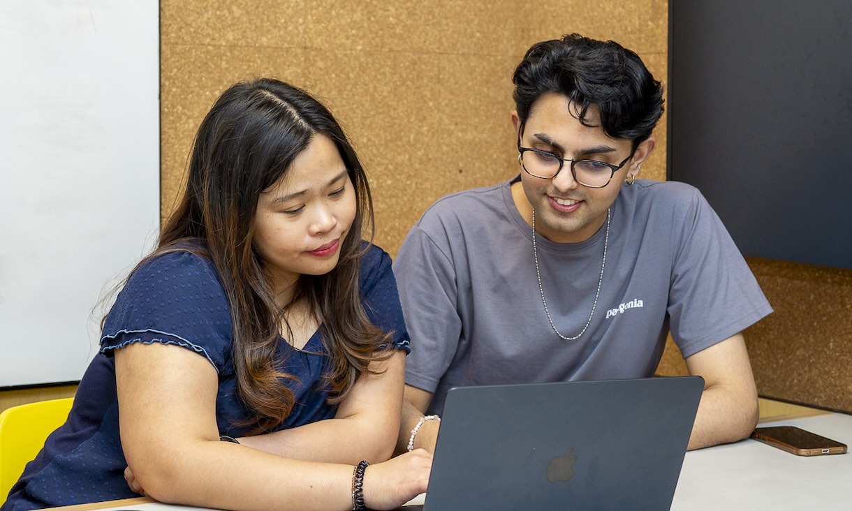 Two students sit together at a table, working on a laptop and discussing their work in a classroom or study area..