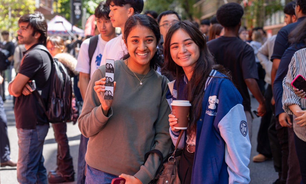 Two students stand on Bowen Street at RMIT University during O Week Semester 1, holding a photo strip and a takeaway coffee, with a crowd and event stalls in the background.