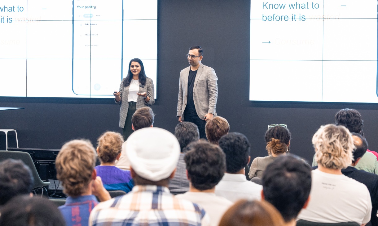 Two presenters stand at the front of a room speaking to a seated audience, with startup presentation slides displayed on large screens behind them at RMIT Activator.