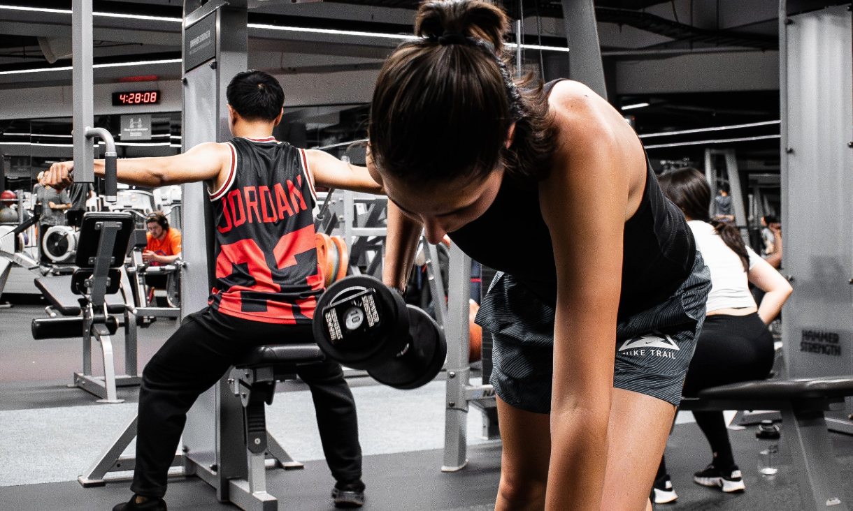 People using strength training equipment in a gym, including free weights and seated cable machines.