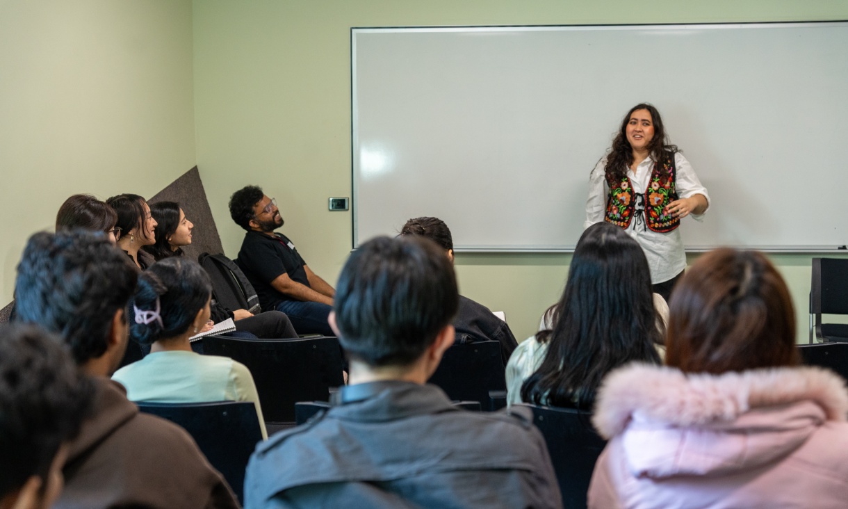 People sit around a person delivering a speech in a classroom setting.