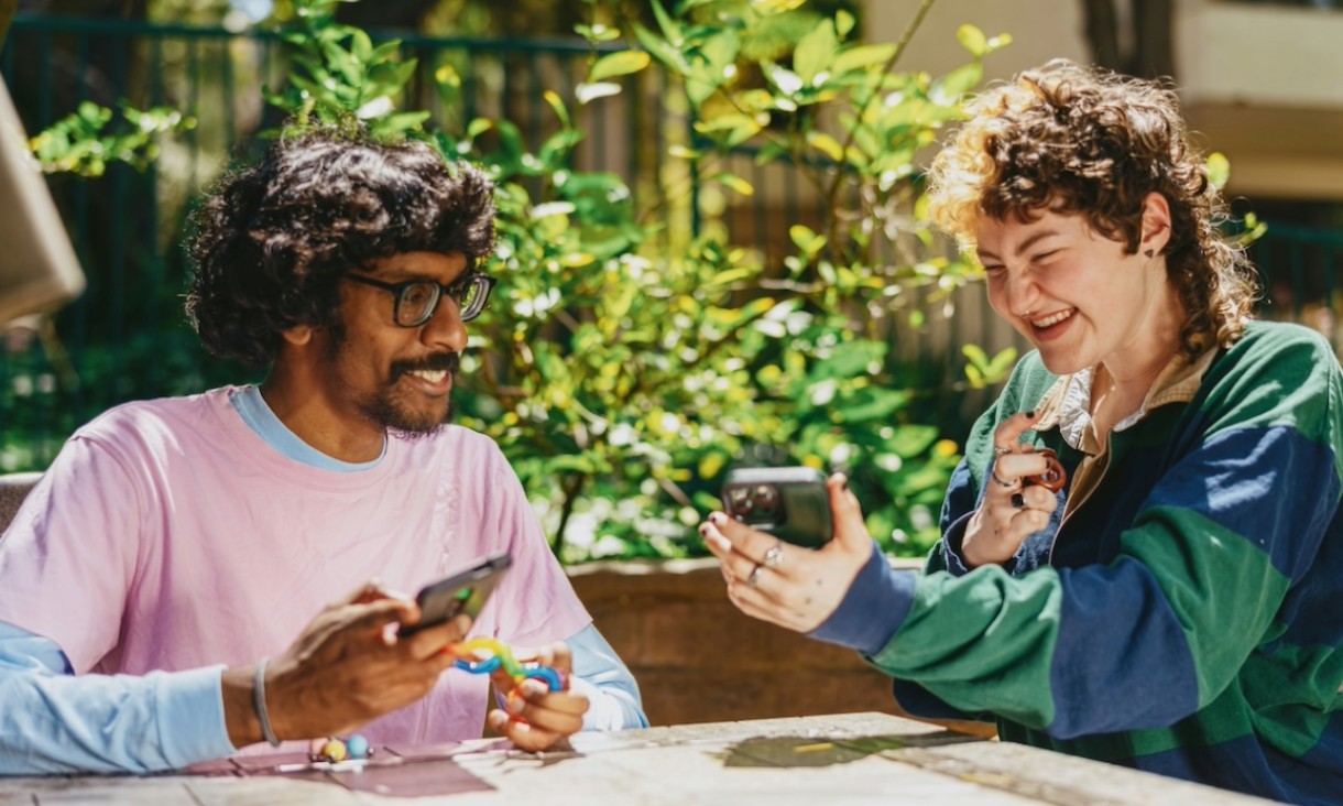 Two people sitting together on a park bench outdoors looking down at a phone together.