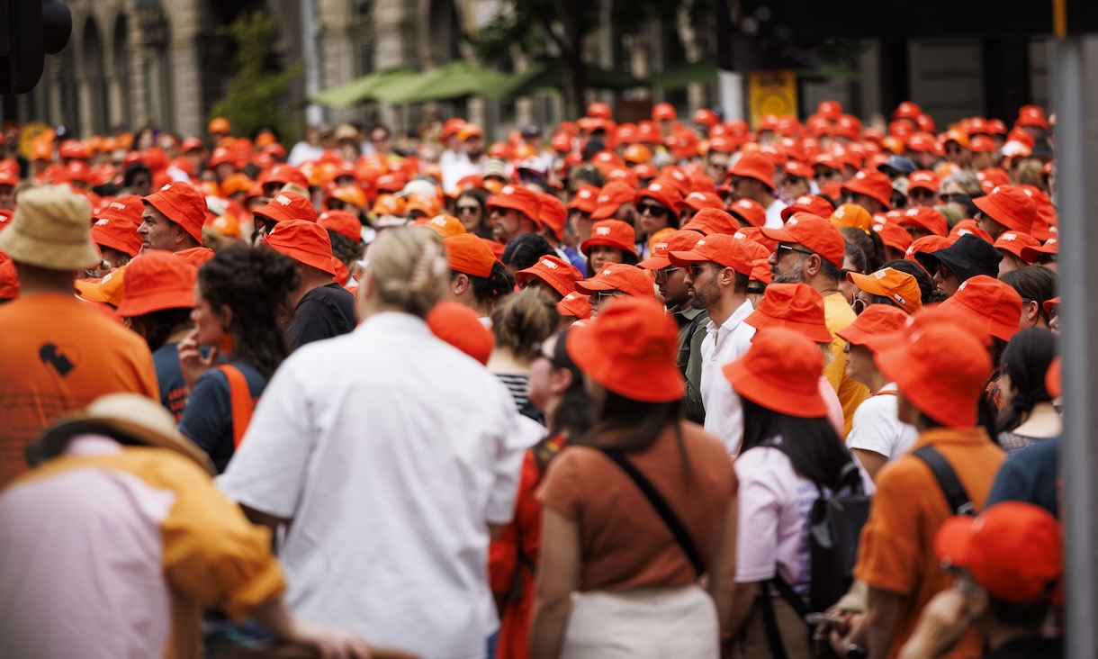 A large crowd of people gathers outdoors, most wearing bright orange hats and shirts, creating a sea of orange at what appears to be a public event or rally.