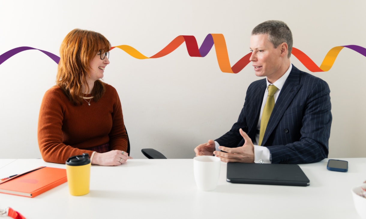 Two people seated across a table in a workplace setting, engaged in a conversation. A laptop, notebook and coffee cups are on the table, with a colourful ribbon graphic displayed on the wall behind them.