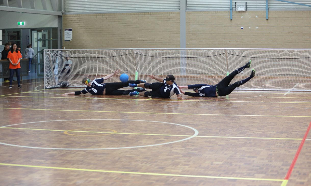 Three goalball players wearing blackout eyeshades dive across an indoor court to block a blue ball in front of the goal.