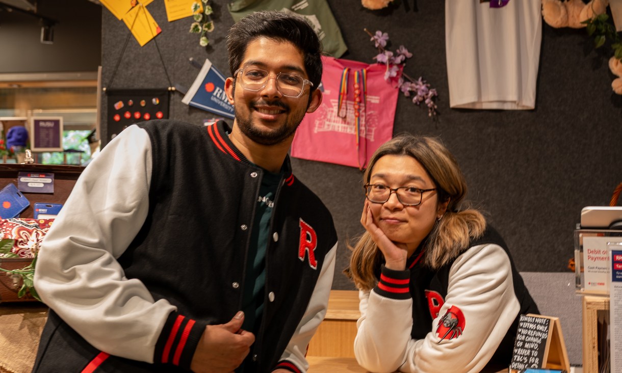 Two people wearing RMIT varsity jackets inside the RMIT Store, featuring black and white panels with red trim and embroidered RMIT detailing.