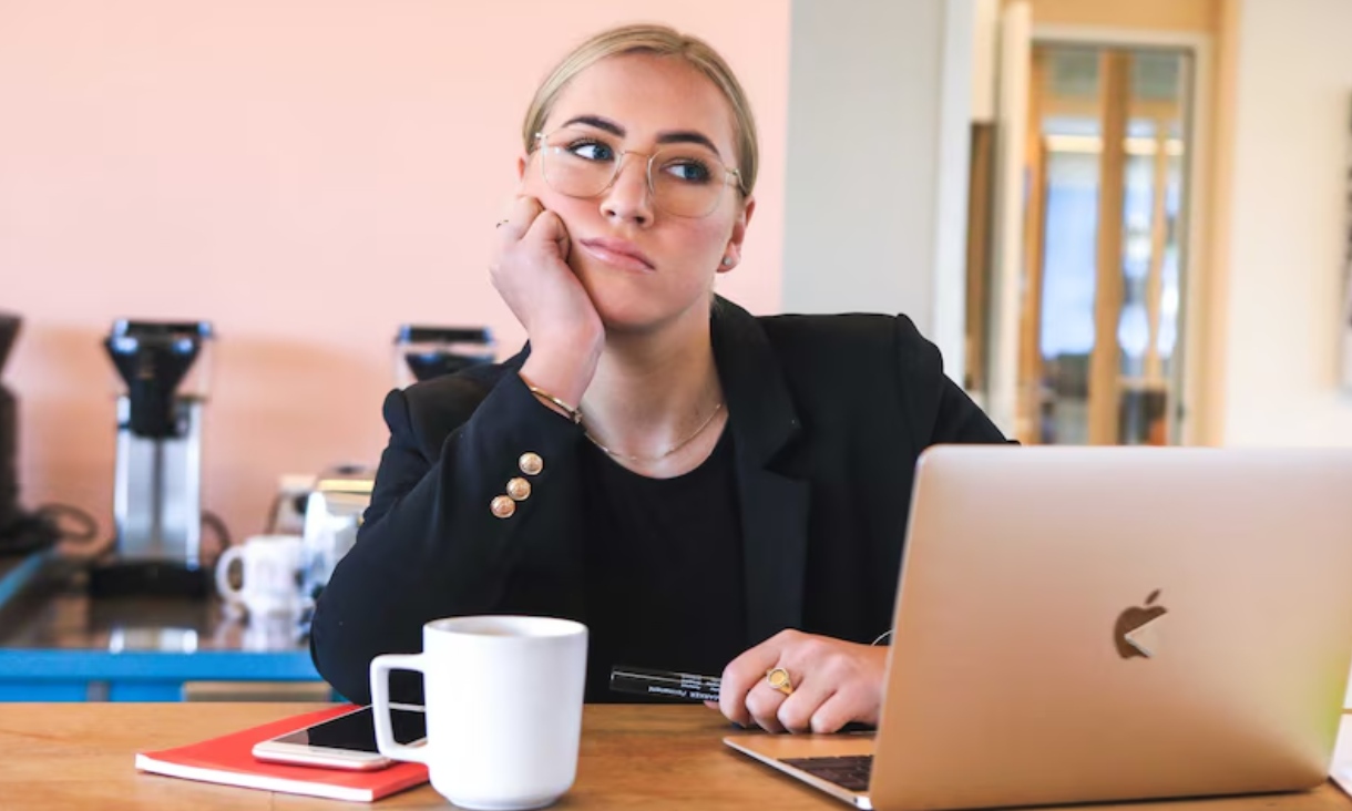 Person resting their chin on one hand while sitting at a table with a laptop, notebook, phone and mug.