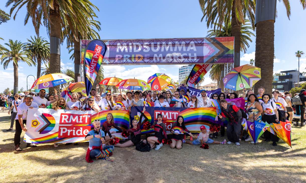 RMIT students, staff and alumni gather beneath the Midsumma Pride March banner in St Kilda, holding RMIT Pride banners, rainbow flags and colourful umbrellas after the march.