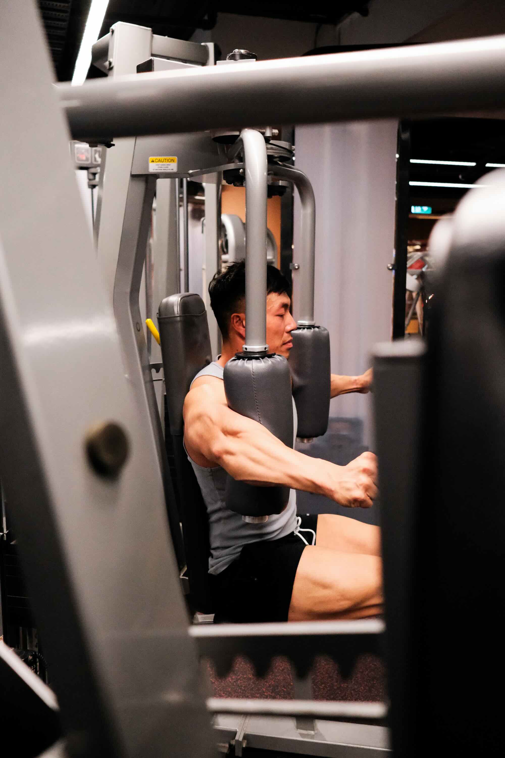 Person using a chest press machine at the gym, focusing on upper body strength training. Their face is not visible, and the scene shows gym equipment and muscular arms in action.