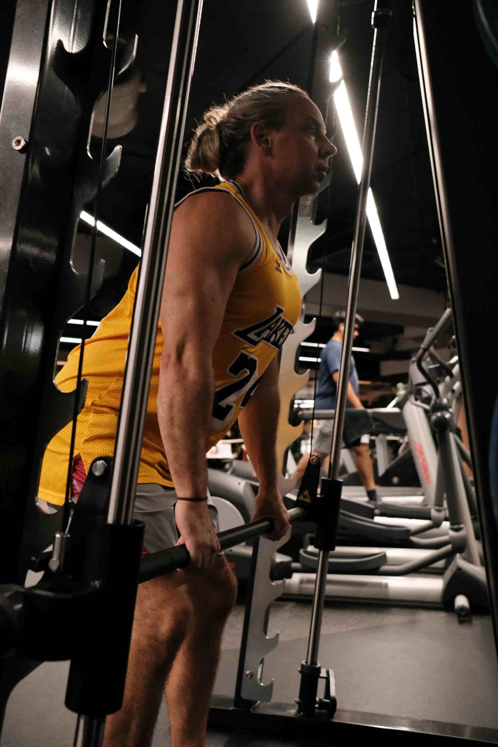 Person in a yellow sports jersey performing a barbell squat using a Smith machine at the gym, focusing on strength training. Gym equipment and other people are visible in the background.
