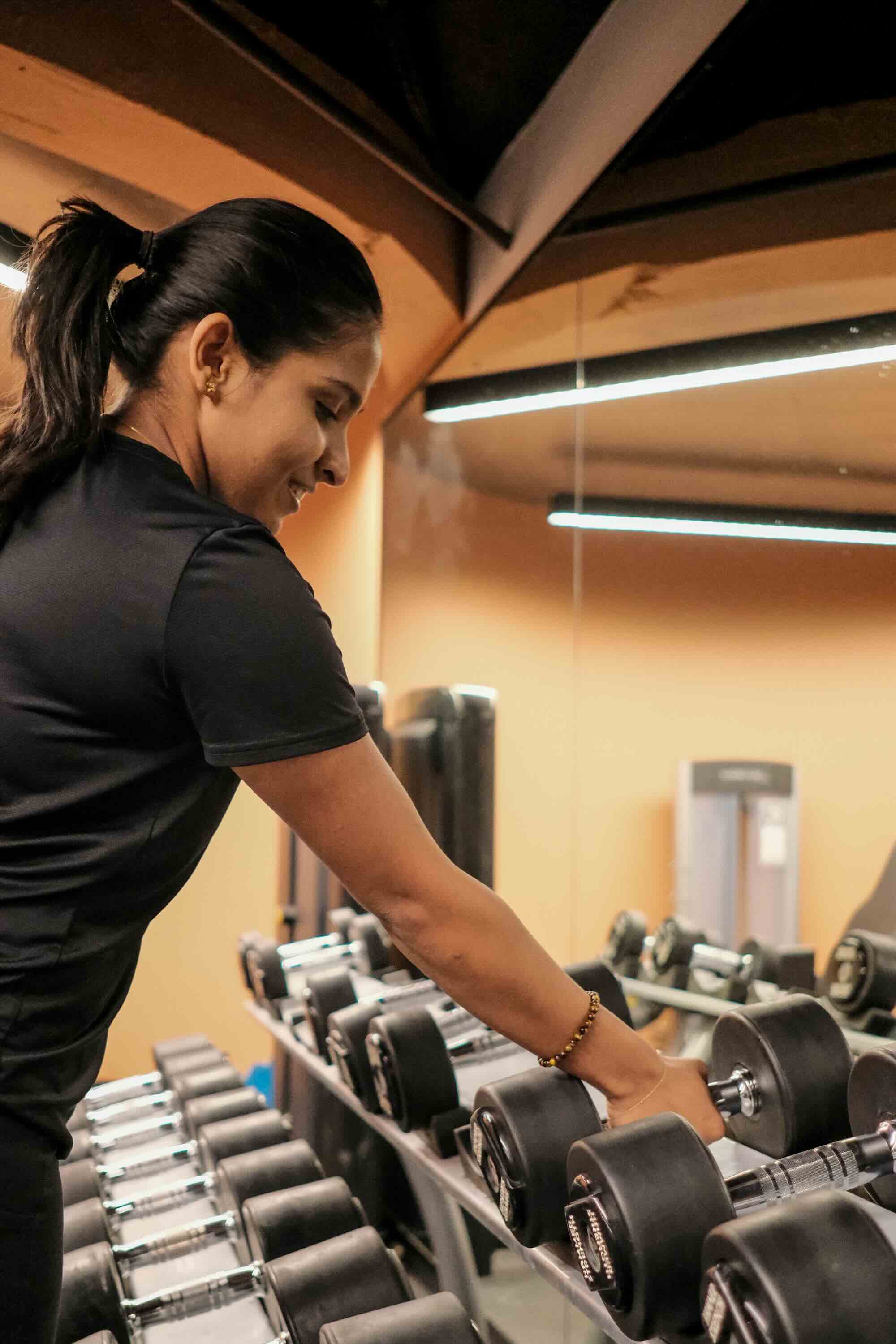 Person in a black shirt selecting a dumbbell from a weight rack in the gym, with a mirrored wall and modern gym equipment visible in the background.
