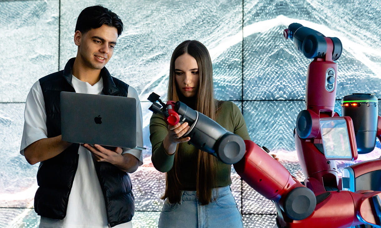 Students interact with a red robotic arm in a laboratory setting, holding a laptop and remote control, with a large digital display in the background.