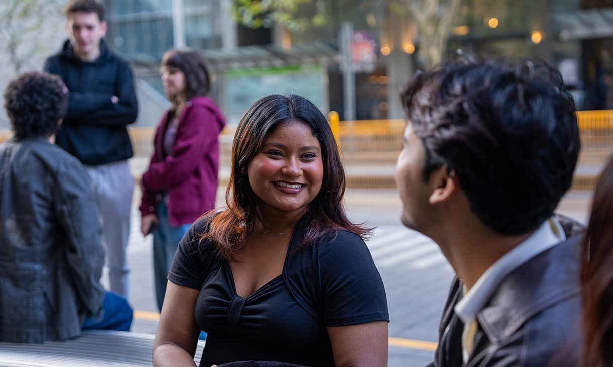 A young woman smiling and talking with a friend outdoors on campus, with other students in the background engaging in conversation.