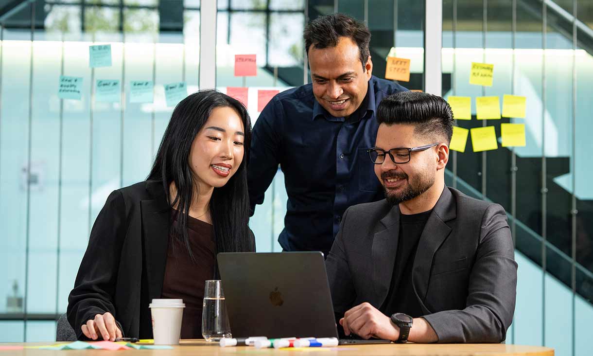 Three professionals working together at a table with a laptop, drinks, and sticky notes, discussing ideas in a modern office.