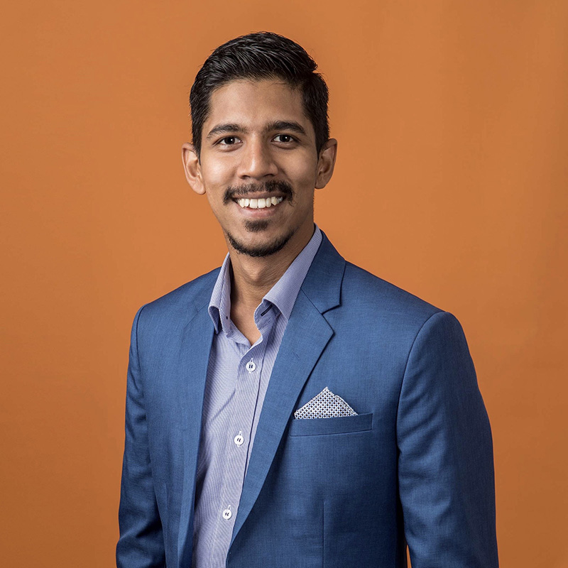 Mevan Peiris in a blue suit and light shirt standing against an orange background. Face is blurred for privacy.