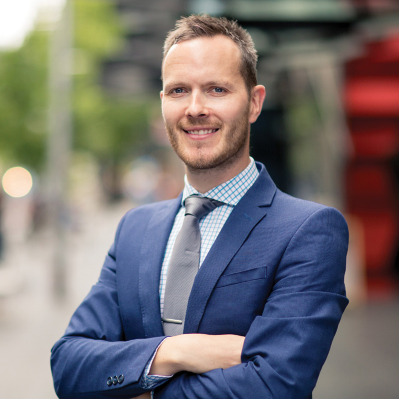 Person in blue suit, checked shirt, and tie with arms crossed, standing outdoors.