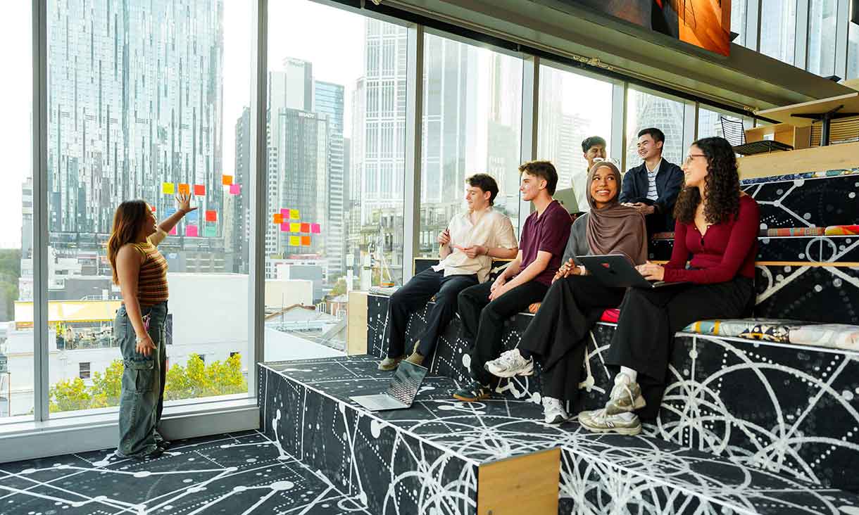 A group of students sit on tiered seating, engaged and smiling, as one student stands by a window covered in colourful sticky notes, presenting to the group in a modern classroom with city views.